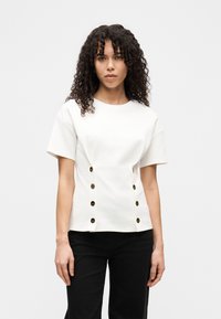 Λευκό τοπ με κοντά μανίκια από απαλό ύφασμα, με στρογγυλή λαιμόκοψη, λεπτομέρειες με κουμπιά στο πλάι και εφαρμοστή γραμμή. Φορεμένο με μαύρο παντελόνι.
