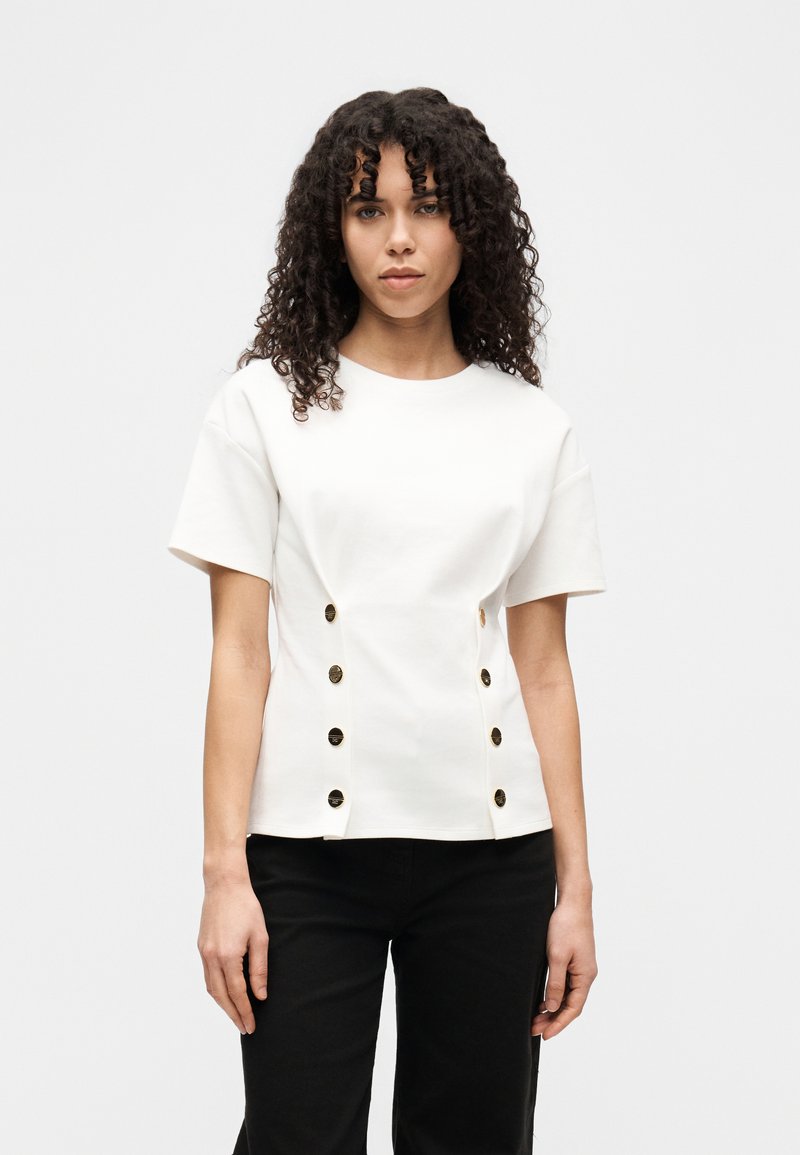 Λευκό τοπ με κοντά μανίκια από απαλό ύφασμα, με στρογγυλή λαιμόκοψη, λεπτομέρειες με κουμπιά στο πλάι και εφαρμοστή γραμμή. Φορεμένο με μαύρο παντελόνι.