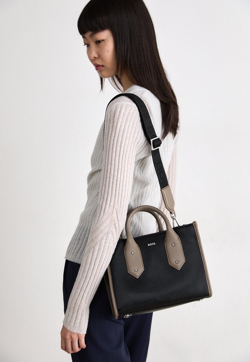 Black leather handbag with beige accents, dual top handles, and adjustable shoulder strap. Features a smooth texture and minimal branding.