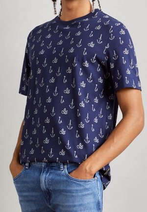Osoba nosząca granatowy t-shirt z białymi wzorami kotwicy i liny, w połączeniu z niebieskimi dżinsami, ręce w kieszeniach, na jednolitym tle.