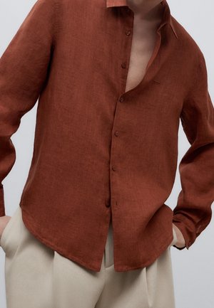 Mężczyzna ubrany w rdzawą lnianą koszulę z guzikami, częściowo rozpiętą, zestawioną z beżowymi plisowanymi spodniami na jednolitym tle.