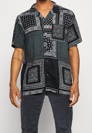 Homme portant une chemise à manches courtes noire et grise à motifs patchwork bandana avec boutons et un pantalon foncé, montrant un tatouage sur l'avant-bras gauche.