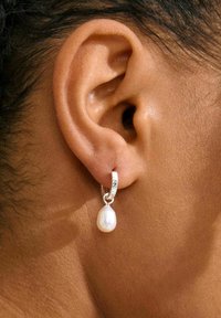 Orecchino a cerchio in argento con una finitura testurizzata, caratterizzato da una singola goccia di perla bianca, indossato sul lobo dell'orecchio su una pelle chiara.