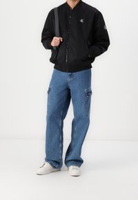 Giacca bomber nera con zip frontale e logo sul petto, abbinata a jeans cargo azzurri chiari con tasche e sneakers bianche.