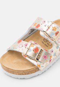 Sandal dengan pola bunga dan tali metalik serta dua gesper emas. Alas kaki tan lembut di atas sol gabus putih, dilengkapi dengan aksen tekstur.