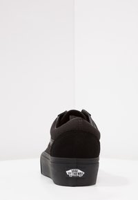 Baskets Vans noires vues du talon montrant le logo "Vans Off The Wall" sur la semelle sur un fond blanc uni.