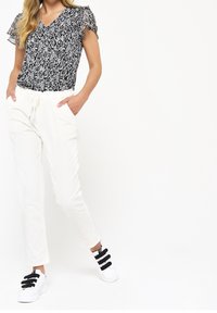 Bílé tepláky se zúženým střihem a kapsami, spárované s černým topem s květinovým vzorem a volánkovými rukávy. Bílé tenisky s černými popruhy.