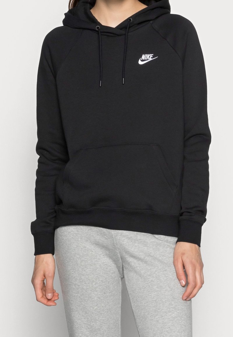 Femme portant un sweat à capuche noir Nike avec poche avant et un pantalon de survêtement gris, debout devant un fond clair uni.