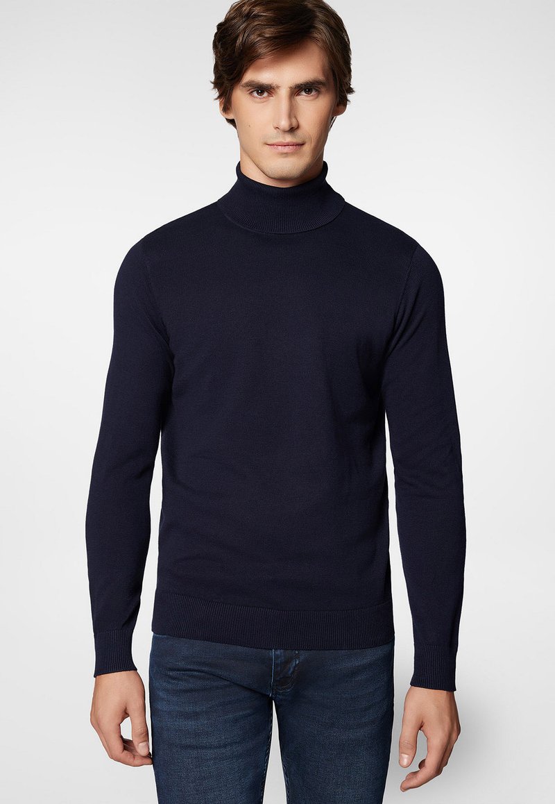 Granatowy sweter typu golf wykonanego z miękkiej dzianiny; długie rękawy; ściągacze przy mankietach i u dołu; noszony z niebieskimi dopasowanymi dżinsami.
