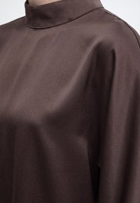 Gros plan sur un tissu lisse brun foncé sur un vêtement à col montant, montrant de doux plis et des détails de couture minimes.