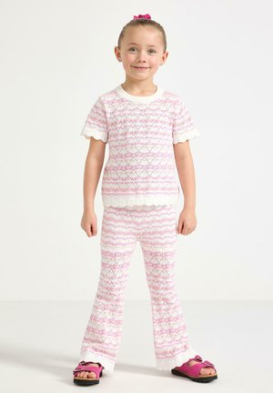 Ung pige står iført matchende lyserødt og hvidt mønstret striktop og bukser med bølgede kanter samt lyserøde sandaler med sløjfer.