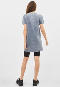 Szara, oversize sukienka t-shirtowa o wyblakłej fakturze, z krótkimi rękawami i zaokrąglonym dołem, połączona z czarnymi szortami rowerowymi i sneakersami.