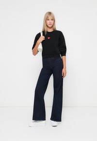 Hanorac negru cu accent în formă de inimă roșie, asortat cu blugi evazați de culoare albastru închis și teneși cu platformă albă. Design simplu și modern.