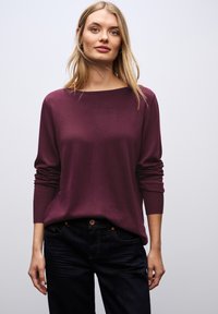 Bordeaux gebreide trui met een bootnek en lange mouwen, gecombineerd met donkere jeans. Gladde textuur, ontspannen pasvorm en geribbelde manchetten.