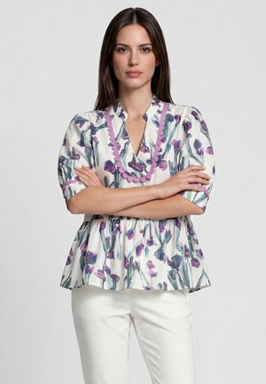 Vrouw met lang bruin haar die een witte blouse met bloemen en paarse accenten draagt en een witte broek, staand met haar armen over elkaar.