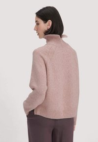 Frau mit kurzem dunklem Haar, die einen hellrosa Rollkragenpullover und dunkle Hosen trägt, in Profil pose mit Händen in den Taschen.