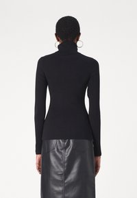 Kvinna med kort hår iklädd en svart ribbad polotröja och svart läderkjol, sedd bakifrån mot en vit bakgrund.