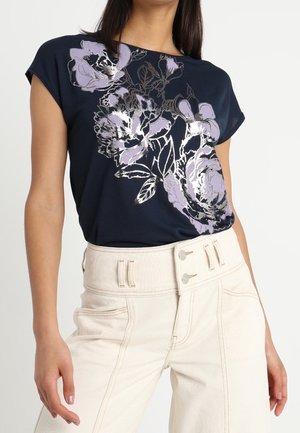 Kobieta ubrana w granatowy top w kwiaty z krótkimi rękawami, włożony w białe spodnie z wysokim stanem i podwójnym zapięciem na guziki.