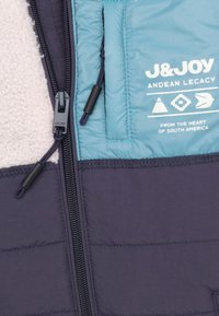 Geïsoleerde jas met een marineblauwe buitenlaag, een lichtblauw paneel en een crèmekleurige fleece voering. Voorzien van een ritsluiting en logo-accenten.