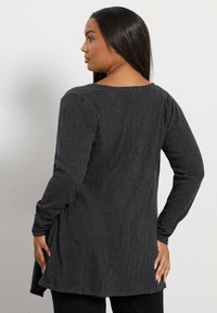 Mörkgrå långärmad tunika med en texturerad tyg, med en vid halsringning och sidofickor. Baksidan har en slät yta.