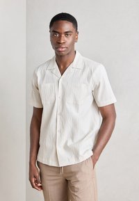 Camisa de manga corta en color crema con textura, dos bolsillos en el pecho y cuello con muesca, combinada con pantalones marrón claro. Acabado de tejido suave.