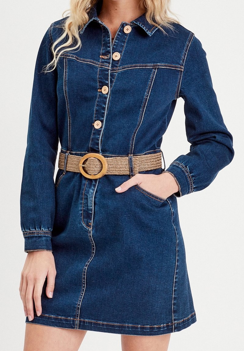 Robe chemise en jean bleu foncé avec des manches longues, boutonnage à l'avant, poche et ceinture à la taille avec une boucle en bois ronde. Coutures contrastantes visibles.