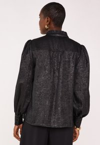 Black textured blouse featuring puffed sleeves, buttoned cuffs, a ruffled collar, and a smooth, slightly shiny finish.
