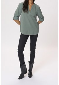 Blusa verde chiaro con colletto e maniche arrotolate, abbinata a jeans skinny neri e stivali da cowboy neri a punta, che mettono in risalto il tessuto texturizzato.