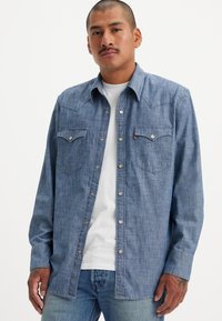 Långärmad denimskjorta i ljusblått, med två spetsiga framfickor och tryckknappar. Stylar en vit t-shirt under.