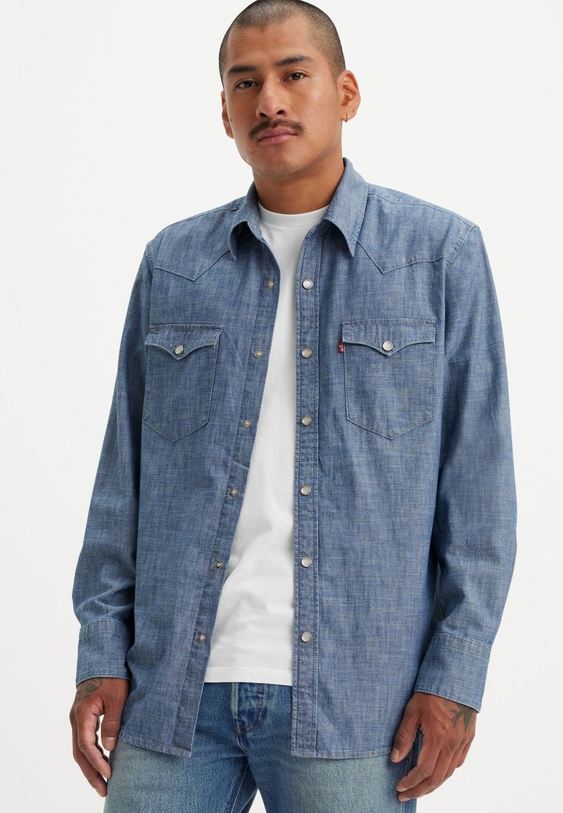 Långärmad denimskjorta i ljusblått, med två spetsiga framfickor och tryckknappar. Stylar en vit t-shirt under.