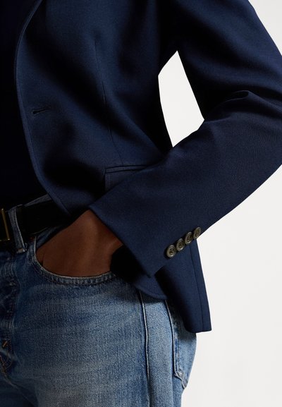 Μπλε σακάκι σε ναυτικό στιλ με εφαρμοστή γραμμή, υφασμένο από υφή με ανάγλυφο ύφασμα, γιακά με εγκοπή και λεπτομέρειες με τέσσερα κουμπιά στα μανίκια, φορεμένο πάνω από ανοιχτόχρωμο τζιν παντελόνι.