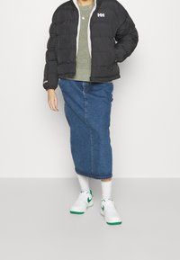 Personne portant une doudoune noire, un pull vert, une jupe midi en denim bleu, des chaussettes blanches et des baskets blanches avec des accents verts, se tenant devant un arrière-plan uni.