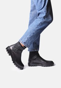 Stivali alla caviglia in gomma nera con finitura testurizzata, caratterizzati da un accento con cinturino e suole robuste e rigate. Indossati con jeans in denim blu.