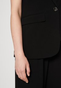Black blazer featuring a single button, side pockets, and structured shoulders; smooth fabric with a matte finish. Hand resting on the fabric.