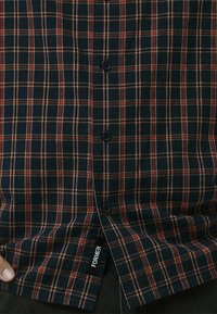 Dark blue plaid shirt with red and cream squares, featuring black buttons and a label on the hem reading "FORMER." Cotton material, smooth texture.