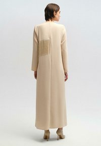 Beige lange jurk met lange mouwen, voorzien van een franjesdetail op de achterkant. Gladde textuur en een rechte silhouette, gecombineerd met beige hakken.