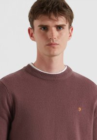 Maglione lavorato a maglia in un profondo colore malva con scollo rotondo e polsini a costine. Presenta un piccolo logo ricamato in arancione a contrasto.