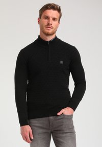 Schwarzer Reißverschluss-Pullover mit einem strukturierten geometrischen Muster, geripptem Kragen und Bündchen, kombiniert mit hellgrauen Jeans. Dezentes Logo-Detail auf der Brust.