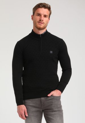Zwarte ritssweater met een gemêleerd geometrisch patroon, geribde kraag en manchetten, gecombineerd met lichtgrijze jeans. Subtiele logo-detail op de borst.