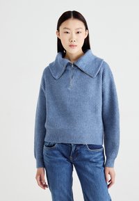 Blå ribbestrikket genser med stor krage og glidelås, kombinert med blå denimjeans. Genseren har lange ermer og en kortere passform.