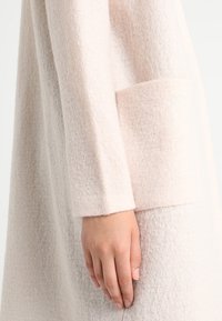 Hellrosa Filzstoffkleid mit strukturierter Oberfläche, langen Ärmeln und einer großen Tasche auf der Vorderseite.