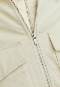 Krēmkrāsas jaka ar metāla rāvējslēdzēju priekšpusē, ar ribotu apkakli un divām kabatām ar atlokiem. Tekstūras audums.