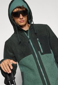 Zeleni i crni zip-up flis džemper s kapuljačom, teksturirani materijal, džep na prsima i podesive vezice. Model nosi usklađeni zimski šešir i sunčane naočale.