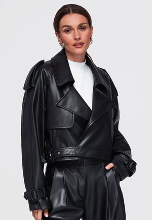 Donna con capelli castani indossa giacca in pelle nera e pantaloni in pelle a vita alta abbinati, posando con una mano in tasca su sfondo semplice.