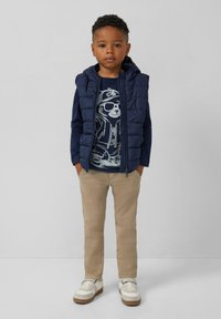 Junger Junge steht mit den Händen in den Taschen, trägt eine marineblaue Pufferweste, ein marineblaues Shirt mit Bärenmotiv, beige Hose und weiße Sneaker.