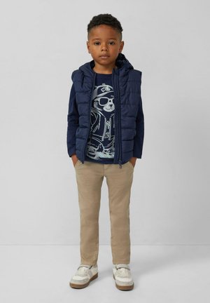 Jongen staat met zijn handen in zijn zakken, draagt een marineblauwe puffervest, een marineblauw shirt met een berenafbeelding, beige broeken en witte sneakers.