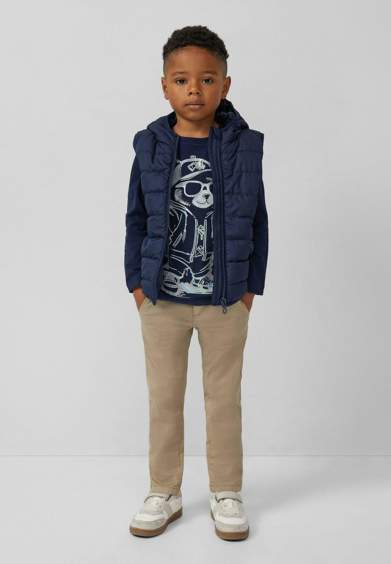 Junger Junge steht mit den Händen in den Taschen, trägt eine marineblaue Pufferweste, ein marineblaues Shirt mit Bärenmotiv, beige Hose und weiße Sneaker.