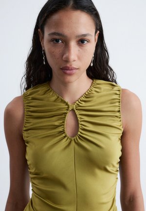 Jonge vrouw met lang donker haar, zilveren oorbellen en een olijfgroene mouwloze top met geplooide details en een sleutelgat-Halslijn.
