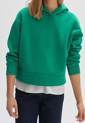 Osoba ubrana w zieloną bluzę z kapturem na białej koszuli i ciemnoniebieskich dżinsach, stojąca z ramionami swobodnie opuszczonymi wzdłuż ciała.