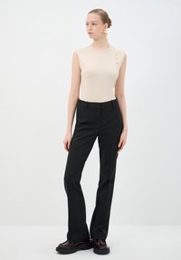 Top sin mangas beige con tres botones dorados en el hombro, combinado con pantalones negros ajustados y zapatos de plataforma burdeos brillantes.